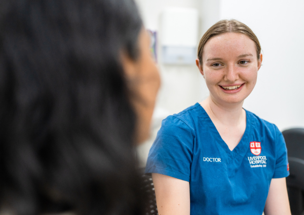 A doctor in blue scrubs smiles and listens while speaking with another person in a clinical setting, showing a calm, respectful conversation.